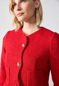Veste texturée rouge avec un col rond, trois fermetures à boutons dorés et deux poches avant avec des détails de couture bien marqués.