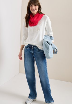 Lachende vrouw die een witte trui, rode sjaal, blauwe spijkerbroek en zilveren sneakers draagt, met een lichtblauwe spijkerjack over haar arm tegen een effen achtergrond.