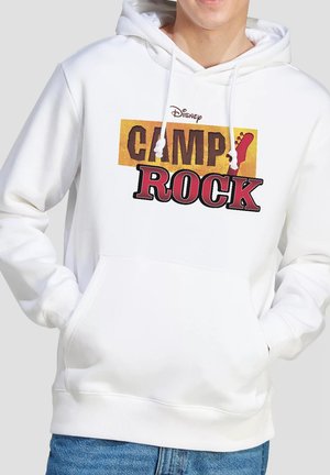 Person wearing white hoodie with "Disney Camp Rock" logo and red guitar headstock graphic, hands in front pocket, blue jeans visible.