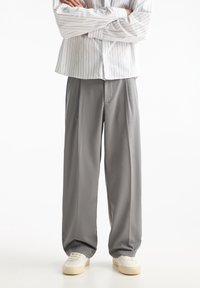 Person stands with arms crossed wearing gray wide-leg trousers, white sneakers, and a white button-up shirt with thin vertical stripes.