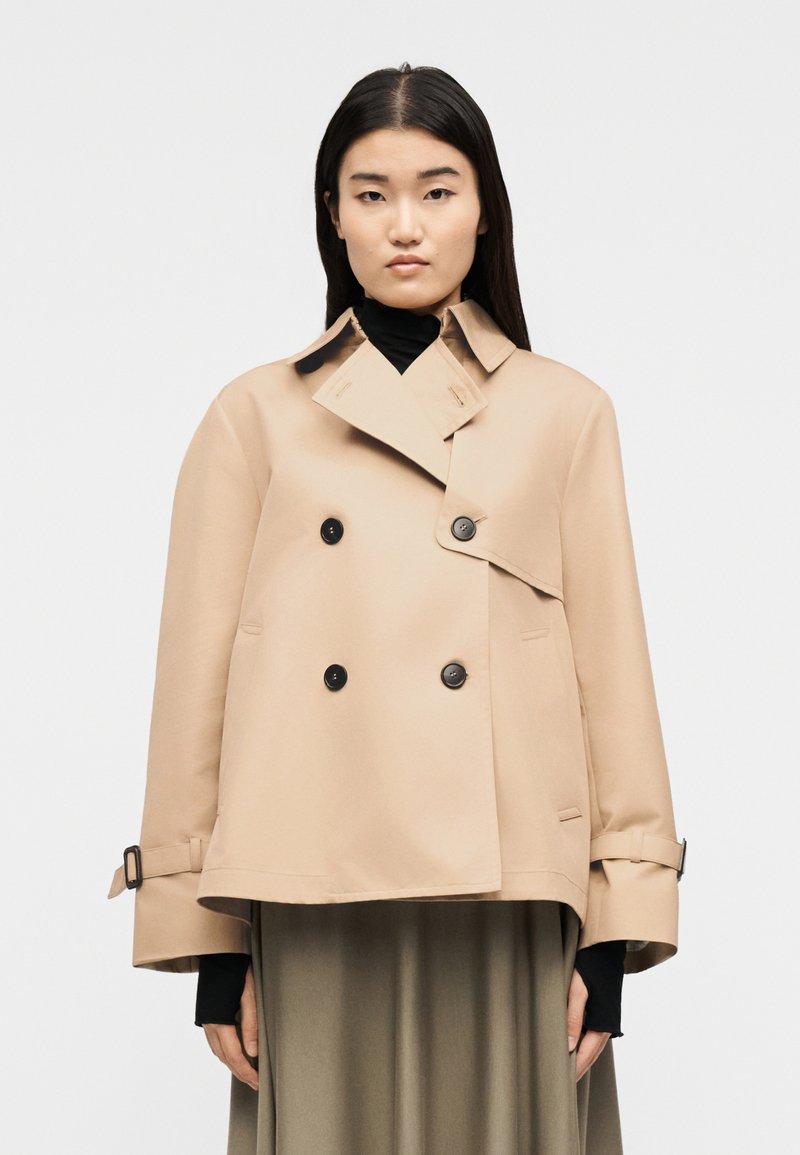 Γυναίκα με μακριά σκούρα μαλλιά, φορώντας μια μπ εζ διπλόκλειστη καμπαρντίνα πάνω από μαύρη ζιβάγκο και ελιάς φούστα, με φόντο λευκό.