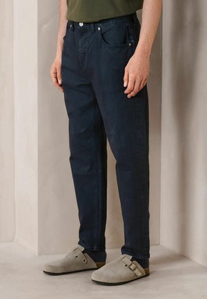 Mann trägt dunkelblaue Jeans, olivgrünes Hemd, schwarze Socken und beige Wildleder-Slipper mit Schnallenriemen, steht vor einer beigen Wand.