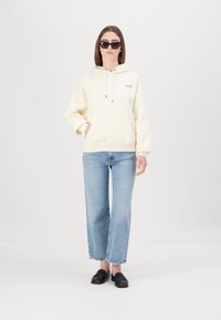 Gaiši dzeltena kapuce ar auklu, priekšējā kabata; apvienota ar gaiši ziliem platām biksēm un melniem loafers, modelis valkā tumšas saulesbrilles.