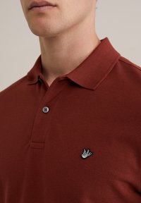 Mørkerød polo skjorte med tekstureret stof, der har spids krave, to knapper og et broderet fuglelogo på venstre bryst.