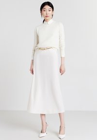 Cremefarbener Pullover, weißes Hemd mit Kragen, fließender weißer Rock. Ein goldener Gürtel betont die Taille. Spitz zulaufende weiße Absatzschuhe vervollständigen den Look.