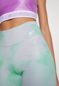 Legginsy Nike w jasnoszarym kolorze z zielonym wzorem tie-dye. Wysoki stan, gładki materiał i małe białe logo na pasie.