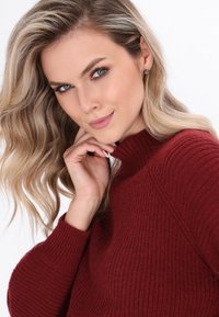 Bordowy sweter robiony na drutach z prążkowaną fakturą i wysokim kołnierzem. Modelka ma faliste blond włosy i opiera brodę na dłoni.
