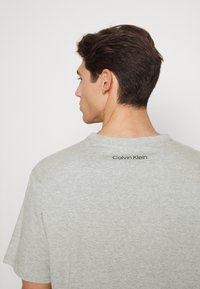 Pilkas trumpomis rankovėmis marškinėliai, pagaminti iš minkštos medžiagos. Yra apvalus kaklo iškirpimas ir juodas "Calvin Klein" logotipas ant viršutinės nugaros dalies.