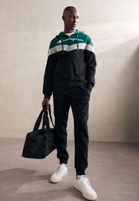 Sudadera con cremallera negra y verde con una franja gris, pantalones negros a juego y zapatillas blancas. Llevando un bolso de viaje negro, de pie sobre una superficie clara.