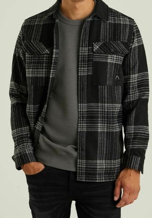 Man wearing black and gray plaid button-up shirt over a gray ribbed sweater, with one hand in pocket and black pants.