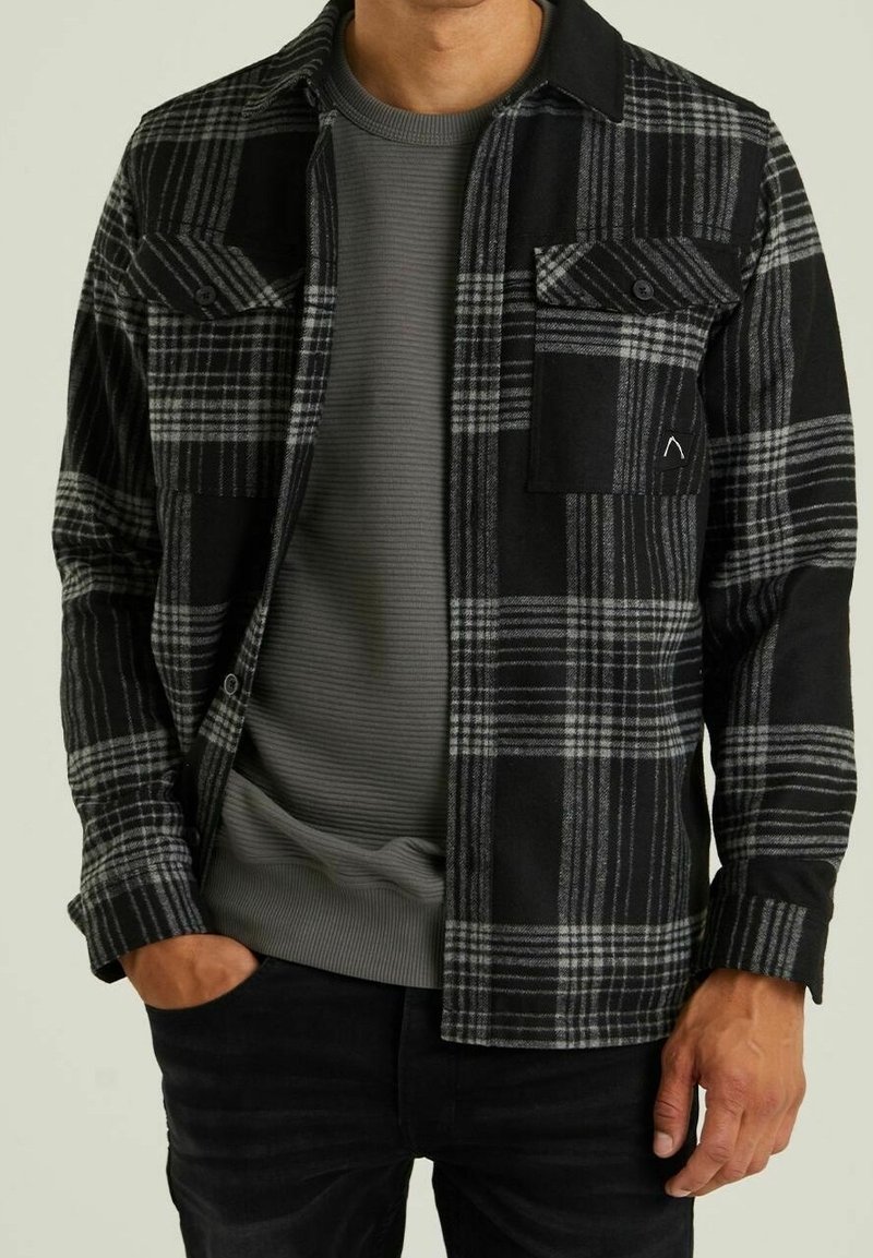 Homme portant une chemise à carreaux noire et grise boutonnée par-dessus un pull côtelé gris, avec une main dans la poche et un pantalon noir.