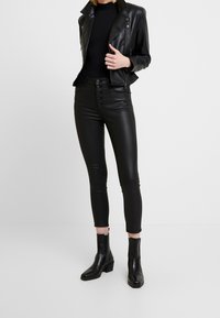 Černá kožená bunda s vypasovaným designem, spárovaná s černými vysokými kalhotami z umělé kůže, které mají pět knoflíků, a kotníkovými botami.