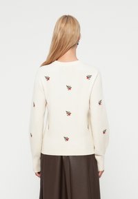 Κρεμ πουλοβεράκι πλεκτό με κεντητά κόκκινα λουλούδια και πράσινα φύλλα· διαθέτει στρογγυλή λαιμόκοψη και μανσέτες και τελείωμα με ριμπ ύφανση.