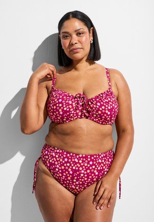 Frau mit schulterlangem schwarzen Haar, die einen pinken, geblümten Bikini trägt, steht vor einer weißen Wand mit Schatten hinter ihr.