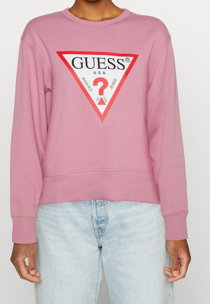 Sweat-shirt rose arborant un grand logo triangulaire Guess en rouge et blanc. Poignets et ourlet côtelés, coupe décontractée avec légère élasticité.