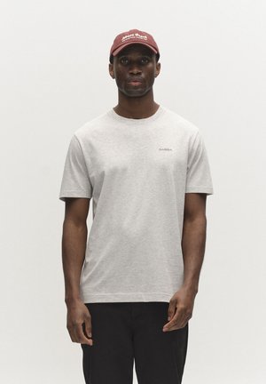 Bărbat purtând o cămașă cu mâneci scurte de culoare gri deschis cu logo mic „GABBA”, pantaloni negri și o șapcă de baseball bordo cu text alb, stând în fața unui fundal simplu.