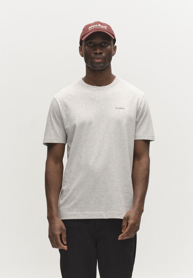 Uomo con maglietta a maniche corte grigio chiaro con piccolo logo "GABBA", pantaloni neri e cappellino da baseball bordeaux con scritta bianca, in piedi davanti a uno sfondo semplice.