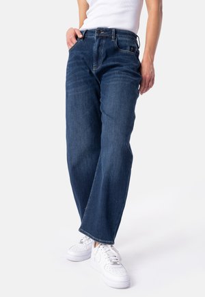 Person trägt dunkelblaue weite Jeans, weiße Sneakers und ein weißes ärmelloses Oberteil, steht mit einer Hand in der Tasche.