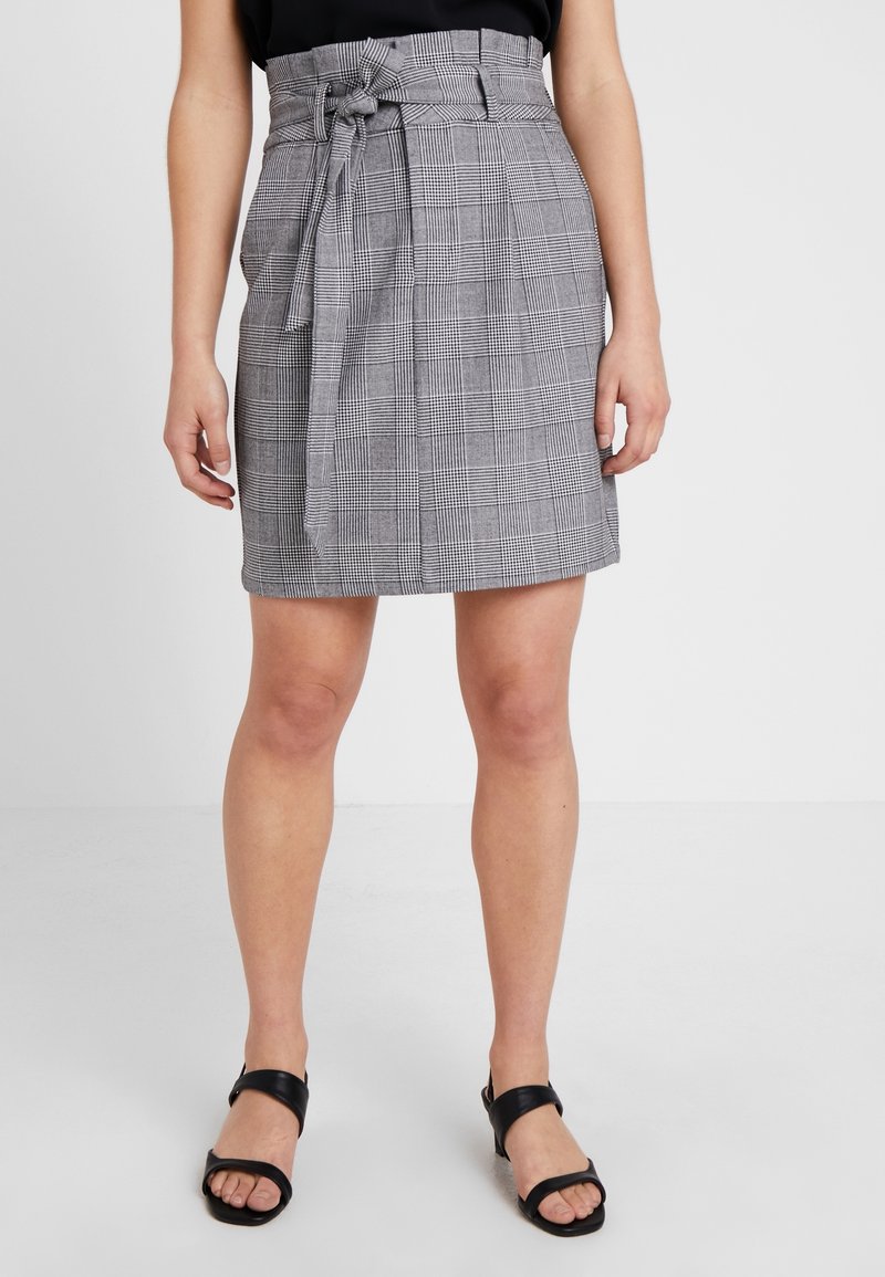 Femme portant une jupe grise à carreaux taille haute avec ceinture nouée à l'avant, un haut noir et des sandales noires à bout ouvert, debout sur un sol blanc.