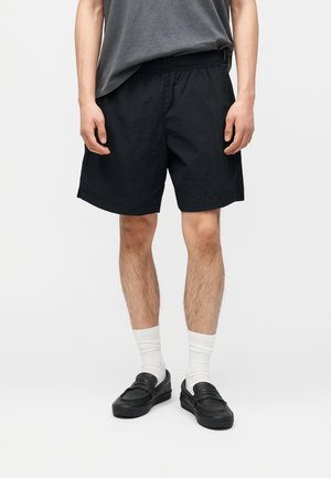 Mężczyzna w czarnych szortach, białych skarpetkach do kostek, czarnych butach wsuwanych i szarej koszulce stojący na białym tle.