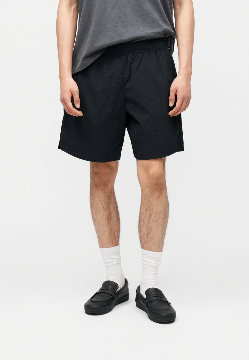 Άνδρας που φοράει μαύρο σορτς, λευκές κάλτσες, μαύρα παπούτσια χωρίς κορδόνια και γκρι μπλουζάκι, στέκεται μπροστά σε λευκό φόντο.