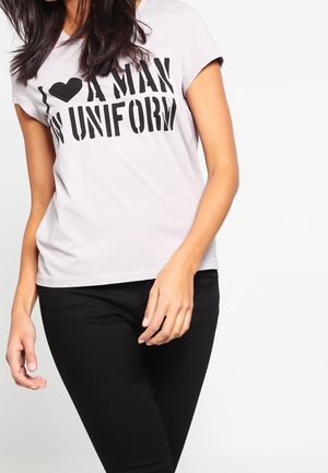 Kvinde iført en hvid t-shirt med teksten "I love a man in uniform" og sorte bukser, stående mod en ensfarvet hvid baggrund.