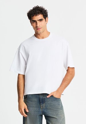 Uomo con capelli ricci che indossa una maglietta bianca larga e jeans blu sbiaditi con una mano in tasca, in piedi davanti a uno sfondo semplice.