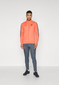 Helle pinke Zip-Jacke mit grauen Akzenten, kombiniert mit grauen Hosen, die pinke Seitestreifen haben. Hergestellt aus glattem, athletischem Stoff.
