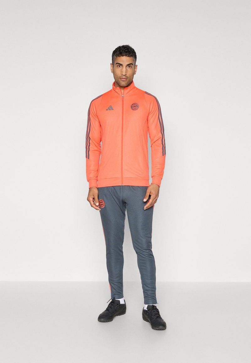 Helle pinke Zip-Jacke mit grauen Akzenten, kombiniert mit grauen Hosen, die pinke Seitestreifen haben. Hergestellt aus glattem, athletischem Stoff.