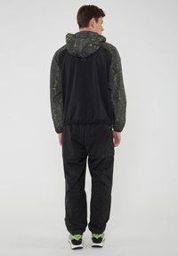 Giacca a vento nera con cappuccio e maniche verde a fantasia, abbinata a pantaloni neri elasticizzati. Presenta polsini elasticizzati e una vestibilità comoda.