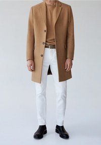 Hombre vestido con abrigo y suéter color camel, pantalones blancos, cinturón marrón y botas de cuero negras hasta el tobillo, de pie contra un fondo plano.
