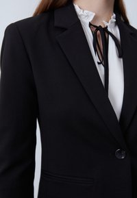 Černý blazer se strukturovaným designem, zapínáním na jeden knoflík a klopami, kombinovaný s bílou halenkou s volánovým límcem.