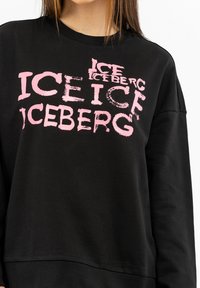Schwarzer Pullover mit großen, sich wiederholenden pinken Texten "ICE" und "ICEBERG" in verschiedenen Schriftarten. Weicher Stoff mit lockerer Passform und langen Ärmeln.