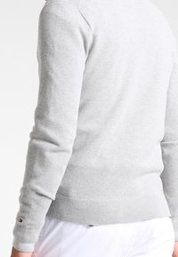 Lichtgrijze gebreide trui met een gestructureerd patroon, geribbelde manchetten en zoom, voorzien van een klein logo-accent vlakbij de manchet.