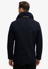 Man met kort bruin haar, gekleed in een marineblauwe wollen jas met opstaande kraag, gezien van achteren tegen een effen witte achtergrond.