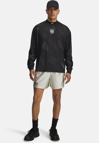 Svart långärmad träningsskjorta med geometriska mönster, kombinerad med ljusa shorts. Inkluderar logotyp och bärs med en svart keps och skor.