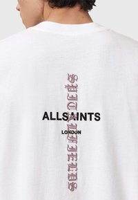 Valkoinen puuvillapaita, jossa on musta "ALLSAINTS LONDON" -logo ja vaaleanpunainen brodeerattu teksti takana. Lyhyet hihat, pyöreä kaula-aukko.