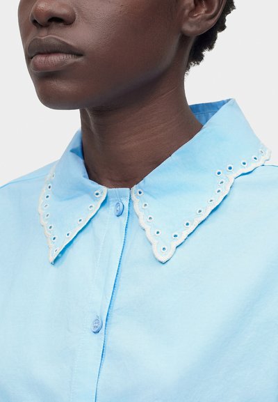 Άτομο με σκουρόχρωμη επιδερμίδα που φοράει γαλάζιο πουκάμισο με κυματιστό, κεντητό γιακά και κουμπωτή μπροστινή πλευρά, σε λευκό φόντο.