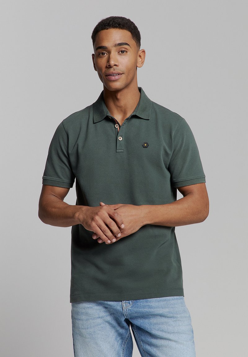Donkergroene polo met een kraag, twee knopen en een klein logo op de borst. G gedragen met lichtblauwe jeans. Gladde katoenen textuur.