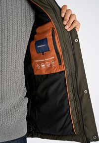 Mørkegrøn jakke med lynlås og orange for. Funktionerne inkluderer et stofmærke og ikoner for funktionelle egenskaber.