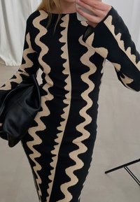 Robe tricotée noire avec des motifs beiges ondulés, manches longues et silhouette ajustée. Les accessoires comprennent un sac à main noir.