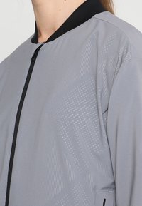 Ljusgrå bomberjacka med dragkedja fram, svart krage och prickig mönsterstruktur, med vinklade designelement längs tyget.