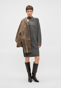 Abito grigio lavorato a maglia, maniche lunghe, collo alto. La modella indossa una giacca di pelle marrone appoggiata su una spalla e stivali marroni fino al ginocchio.