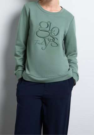 Osoba ubrana w szałwiowo zielony sweatshirt z wypukłym napisem "glovys touch" oraz granatowe spodnie, ręce w kieszeniach na jednolitym tle.