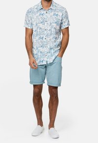 Korte mouw knoopshirt in lichtblauw met een boom- en bergprint; gecombineerd met lichtblauwe opgerolde shorts en witte instappers.