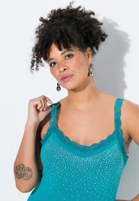 Camiseta sin mangas verde azulado con tirantes de encaje, que presenta un patrón de lunares con tachuelas brillantes en la parte delantera. Textura suave y diseño ajustado.