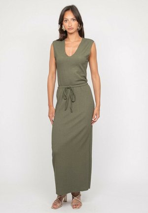 Femme portant une robe longue sans manches vert olive avec un décolleté en V et une ceinture à la taille, associée à des talons beiges à brides et des boucles d'oreilles fleuries.