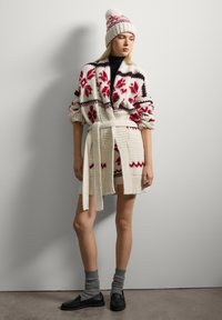 Cardigan in lana con motivi floreali rossi e accenti neri, legato in vita. Abbinato a un cappello coordinato e calze grigie. Mocassini neri completano il look.