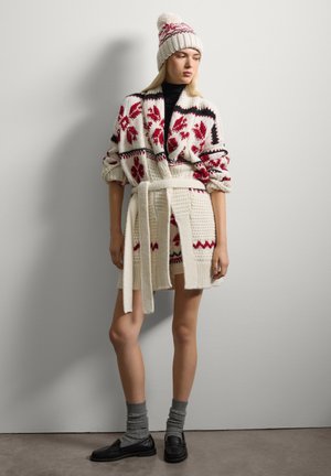 Cardigan in lana con motivi floreali rossi e accenti neri, legato in vita. Abbinato a un cappello coordinato e calze grigie. Mocassini neri completano il look.