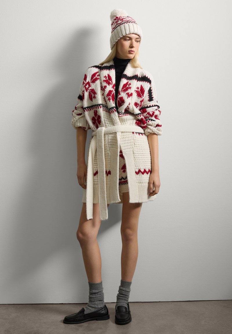 Cardigan in lana con motivi floreali rossi e accenti neri, legato in vita. Abbinato a un cappello coordinato e calze grigie. Mocassini neri completano il look.